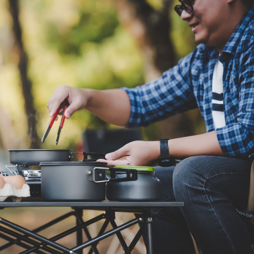 Person cooking outdoors using a portable stove with a blurred natural background

