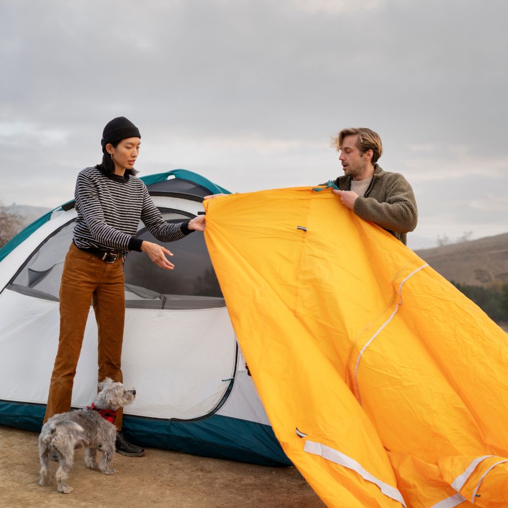 Two people setting up a yellow tent with a dog nearby

