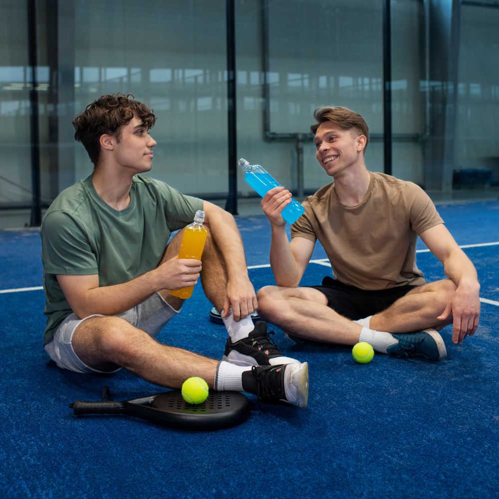 Two young men sitting on a blue court with sports equipment and drinks.

