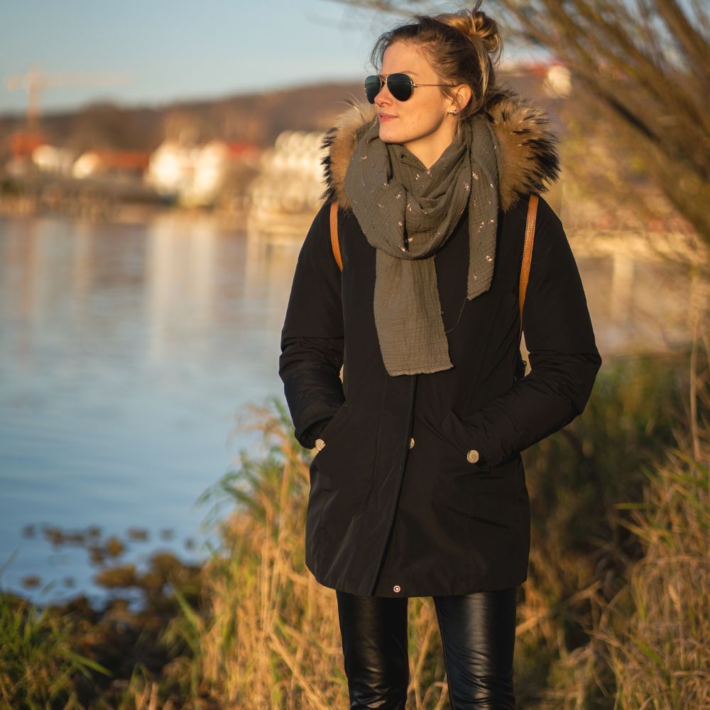 Woman standing by a body of water wearing a black coat with fur hood and sunglasses.

